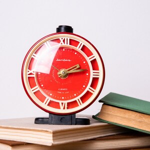 Puede incluir: Un reloj despertador rojo con números romanos en la esfera. El reloj está encima de una pila de libros. La esfera del reloj dice "Junio" y "Libres Made in Japan".