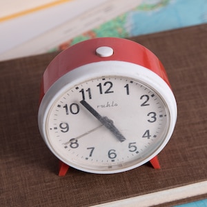 May include: A red alarm clock with a white face and black hands. The clock is set to 10:10 and has the word "Ruhla" printed on the face.