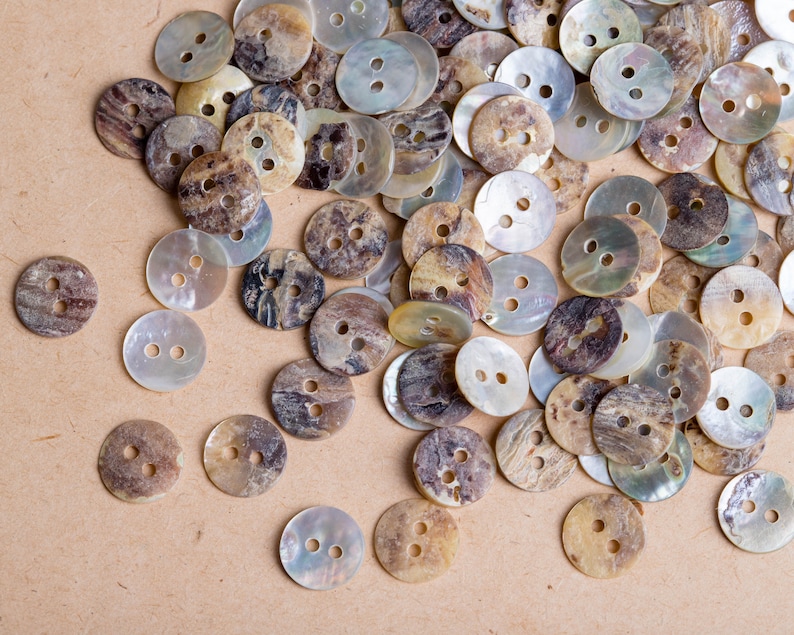 May include: A collection of assorted brown and white shell buttons with a mottled pattern. The buttons are scattered on a light brown surface.