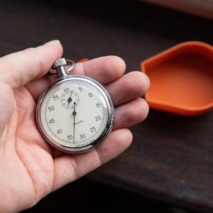May include: A vintage stopwatch with a white face, black numerals, and a chrome case is held in a hand. An orange, teardrop-shaped container sits nearby. The stopwatch is likely used for timing events or measuring intervals.