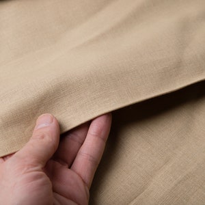 May include: Close-up of tan-colored fabric with a textured weave. The fabric is being held by a hand, showing the material's drape and texture. The neutral color suggests versatility for various sewing or crafting projects.