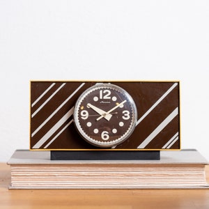 Könnte beinhalten: Eine braun-weiß gestreifte Uhr mit schwarzem Zifferblatt und weißen Zahlen. Die Uhr steht auf einem Stapel Bücher.