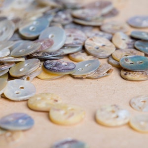 May include: A close-up of a collection of various sized and shaped mother-of-pearl buttons. The buttons are in shades of white, cream, and blue.