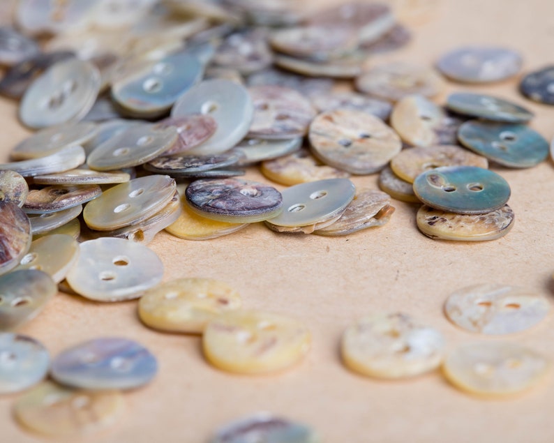 May include: A close-up of a collection of various sized and shaped mother-of-pearl buttons. The buttons are in shades of white, cream, and blue.