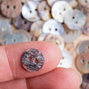 May include: A close-up of a brown and black two-hole button with a mottled pattern. The button is being held by a finger.
