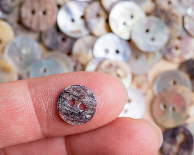May include: A close-up of a brown and black two-hole button with a mottled pattern. The button is being held by a finger.