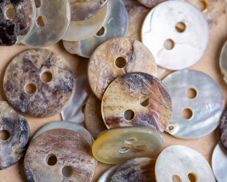 May include: A collection of various sized and patterned buttons made from mother of pearl and other natural materials. The buttons are in shades of brown, white, and cream.