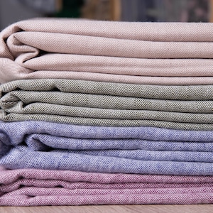 Puede incluir: Cuatro mantas de tela dobladas en diferentes colores: beige, verde, azul y rosa. Las mantas tienen un patrón de espiga y están apiladas una encima de la otra.