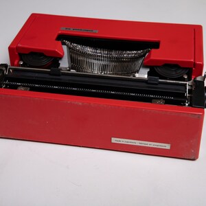 May include: A red Olivetti Lettera 32 typewriter with the lid open, revealing the black keys and the type bars. The typewriter has a black label on the bottom that reads "Made in Yugoslavia - Fabriqu&eacute; en Yougoslavie".