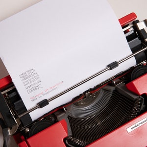 May include: A red Underwood 315 typewriter with a sheet of paper in the carriage. The paper has the numbers 234567890+ and the letters QWERTYUIOPA, qwertyuion, sadfghjkbit, ASDPOHIKTA, sxorbus, and the text "Underwood 315 Typewriter Made in Yougoslavia" printed on it.