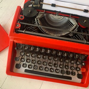 May include: A red vintage typewriter with a black keyboard and a white paper in the carriage. The typewriter has the text "derwood 315" on the side.