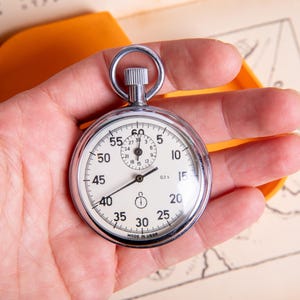 May include: A silver stopwatch with a white face and black markings. The stopwatch has a black second hand and a black minute hand. The face of the stopwatch has numbers from 5 to 60, and the words "0.2 sec" printed on it. The stopwatch is made in the USA.