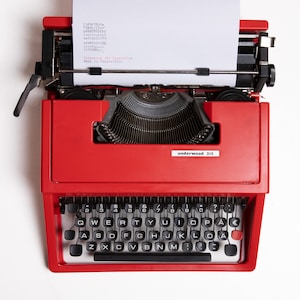 May include: A red vintage Underwood 315 typewriter with a blank sheet of paper inserted. The typewriter has a black keyboard with white keys and a black carriage return lever.