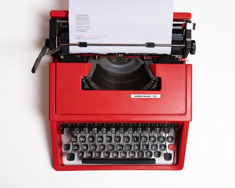 May include: A red vintage Underwood 315 typewriter with a blank sheet of paper inserted. The typewriter has a black keyboard with white keys and a black carriage return lever.