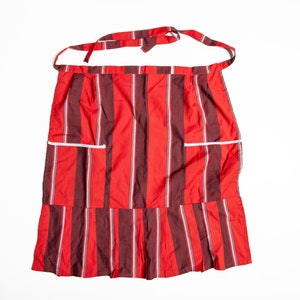 Può includere: Un grembiule a righe rosse e marroni con bordi bianchi e due tasche. Il grembiule ha un fondo arricciato.