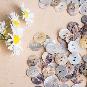 May include: A close-up of a collection of round, iridescent, mother-of-pearl buttons with brown and white marbling. The buttons are arranged in a random pattern on a light brown surface.