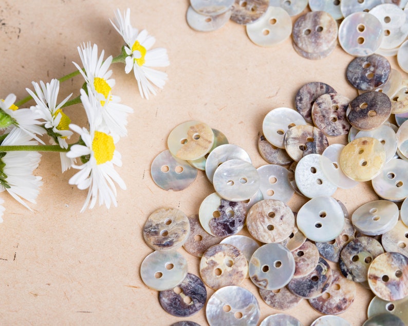 May include: A close-up of a collection of round, iridescent, mother-of-pearl buttons with brown and white marbling. The buttons are arranged in a random pattern on a light brown surface.