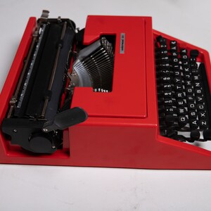 May include: A red vintage typewriter with a black keyboard and a black carriage return lever. The typewriter has a black ribbon and a white label that reads "Olivetti Lettera 32".