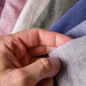 May include: A hand holding a stack of fabric swatches in various shades of grey, blue, and pink. The swatches are made of a soft, textured material.