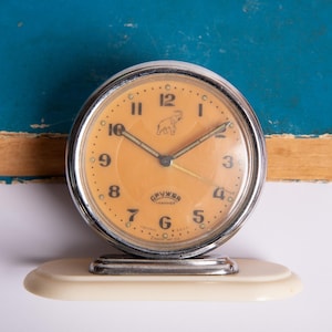 May include: A vintage silver alarm clock with an orange face and black numbers. The clock has a small elephant logo and the text "СГВЧКР" in Cyrillic script. The clock is sitting on a white base.