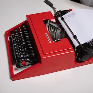 May include: A red Olivetti Lettera 35 typewriter with a black keyboard and a sheet of paper in the carriage. The typewriter has a black lever on the left side and a black knob on the right side.