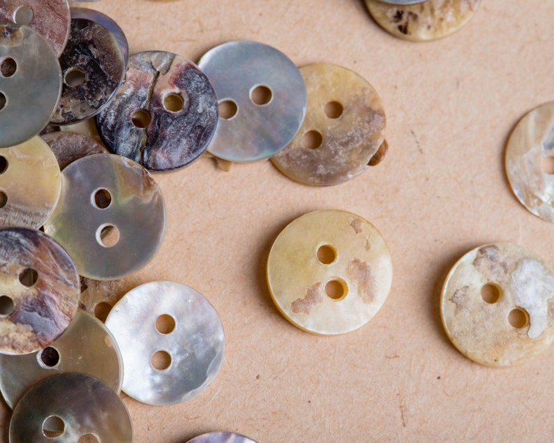 May include: A collection of various sized, two-hole, mother-of-pearl buttons in shades of brown, beige, and white. The buttons have a smooth, shiny finish.