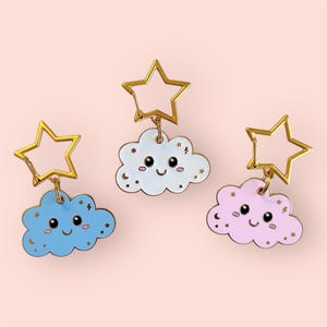 May include: Three cloud-shaped keychains in blue, white, and pink, each with a smiling face and star accents. They hang from gold star-shaped clasps. The background is a soft pink.