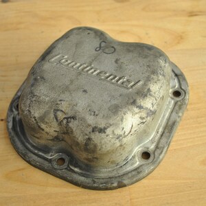 May include: A silver-coloured metal valve cover with the word "Continental" embossed on the top. The cover has a worn, aged appearance.