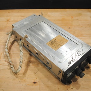 May include: A rectangular, silver-colored electronic device with a black front panel featuring knobs and labels. The device has a label on top and a bundle of white wires. The numbers "13-294" are visible on the side.