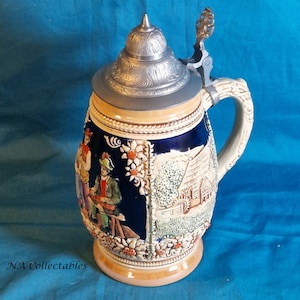 Peut inclure: Une chope à bière en céramique avec un couvercle en étain et une poignée. La chope est décorée d'une scène de village en bleu et blanc avec des personnes et des fleurs.