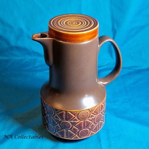 Puede incluir: Una cafetera de cerámica marrón con una tapa marrón y naranja. La cafetera tiene un patrón geométrico en la parte inferior.