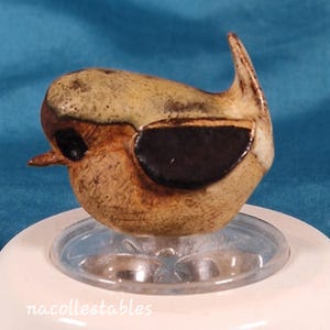 May include: A ceramic bird figurine with a speckled tan and brown body, a dark eye, and a curved tail. The bird is mounted on a clear plastic base and a white surface. The text "nacollectables" is visible.