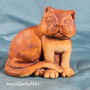 Puede incluir: Figura de gato de cerámica marrón en posición sentada. El gato tiene ojos grandes y redondos y una cara detallada. La figura es de un tono cálido y terroso, con textura y detalles visibles. El texto "nacollectables" es visible.