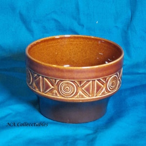 Puede incluir: Un cuenco de cerámica marrón con un patrón geométrico alrededor del borde. El cuenco tiene una base ancha y redonda y un labio ligeramente acampanado.