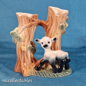 May include: A ceramic vase shaped like two tree trunks with a lamb standing between them. The vase is brown, green and white with black accents.
