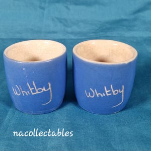 Könnte beinhalten: Zwei blaue Keramikbecher mit dem Wort "Whitby" in weißer Schrift. Die Becher haben einen hellbeigen Innenraum und stehen vor einem türkisblauen Hintergrund. Der Text "nacollectables" steht unten.