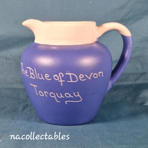 Może przedstawiać: Niebiesko-biały ceramiczny dzbanek z uchwytem. Na dzbanku widnieje napis "The Blue of Devon Torquay" białym pismem. Uchwyt i górna krawędź są białe. Przedmiot kolekcjonerski.