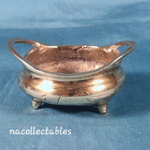 Puede incluir: Un pequeño cuenco de metal plateado con tres patas cortas y dos asas curvas. El cuenco tiene una banda decorativa alrededor del centro y un interior ligeramente texturizado. El fondo es de color azul liso.