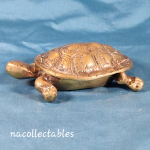 Peut inclure: Figurine de tortue en laiton détaillée, avec une carapace texturée et des membres tendus. La tortue est de couleur or chaud et repose sur une surface bleue. La figurine est un objet décoratif.