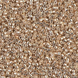 May include: A close-up shot of a pile of small, metallic gold-colored seed beads. The beads are cylindrical with rounded edges and small holes, creating a textured surface. The beads are scattered, filling the frame.