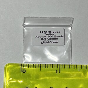 May include: A clear plastic bag containing 11/0 Miyuki Delica beads. The bag is labelled with the text "11/0 Miyuki Delica Approx 500 Beads 2.5 Grams Ja-Pette Beads". The bag is sitting on a yellow ruler.