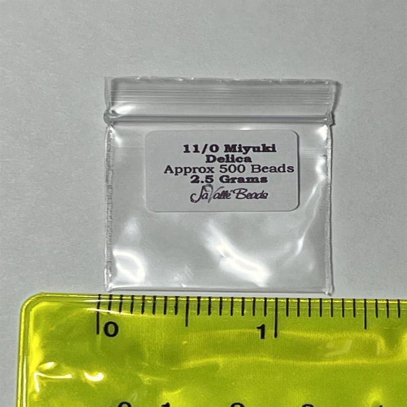 May include: A clear plastic bag containing 11/0 Miyuki Delica beads. The bag is labelled with the text "11/0 Miyuki Delica Approx 500 Beads 2.5 Grams Ja-Pette Beads". The bag is sitting on a yellow ruler.