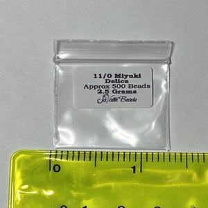 Pu&ograve; includere: Un sacchetto di plastica trasparente contenente perline Miyuki Delica 11/0. Il sacchetto &egrave; etichettato con il testo "11/0 Miyuki Delica Approx 500 Beads 2.5 Grams Ja-Pette Beads". Il sacchetto &egrave; appoggiato su un righello giallo.