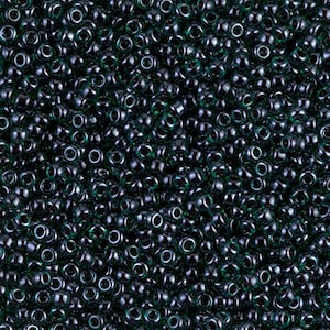 May include: A close-up view of a large quantity of dark green glass seed beads. The beads are small and round, with a hole in the center. They are arranged in a random pattern, creating a textured surface.