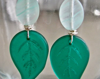 Può includere: Un paio di orecchini fatti a mano con un design a forma di foglia. Ogni orecchino ha un ciondolo a forma di foglia verde acqua con dettagli incisi, collegato a una perla ovale azzurra e un gancio color argento.