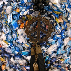 Pu&ograve; includere: Una collana con un ciondolo a forma di segno di pace, perline nere e un fiocco. La collana &egrave; in metallo e ha un aspetto vintage. Lo sfondo &egrave; un tessuto blu e bianco.