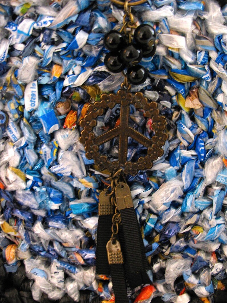 Pu&ograve; includere: Una collana con un ciondolo a forma di segno di pace, perline nere e un fiocco. La collana &egrave; in metallo e ha un aspetto vintage. Lo sfondo &egrave; un tessuto blu e bianco.