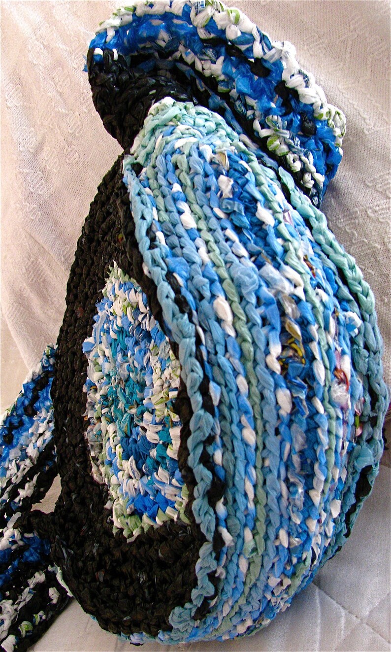 Pu&ograve; includere: Un tappeto all'uncinetto blu e bianco realizzato con sacchetti di plastica riciclati. Il tappeto ha una forma circolare e un bordo nero.