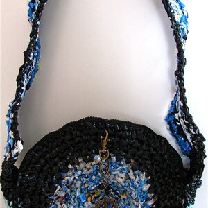 Pu&ograve; includere: Una borsa rotonda in crochet nero e blu con un lungo cinturino. La borsa &egrave; realizzata con materiali riciclati e ha un ciondolo a forma di segno della pace.