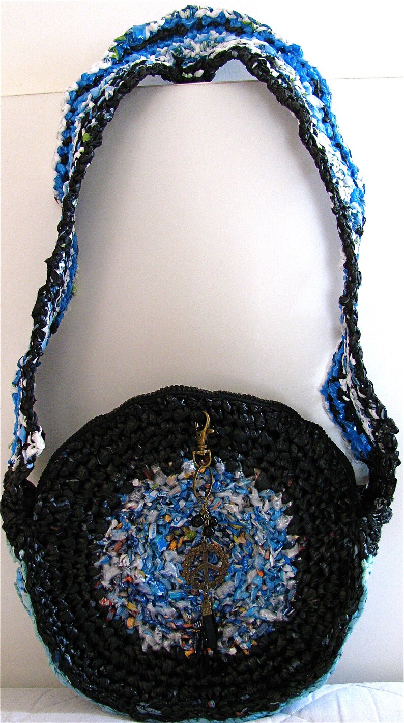 Pu&ograve; includere: Una borsa rotonda in crochet nero e blu con un lungo cinturino. La borsa &egrave; realizzata con materiali riciclati e ha un ciondolo a forma di segno della pace.